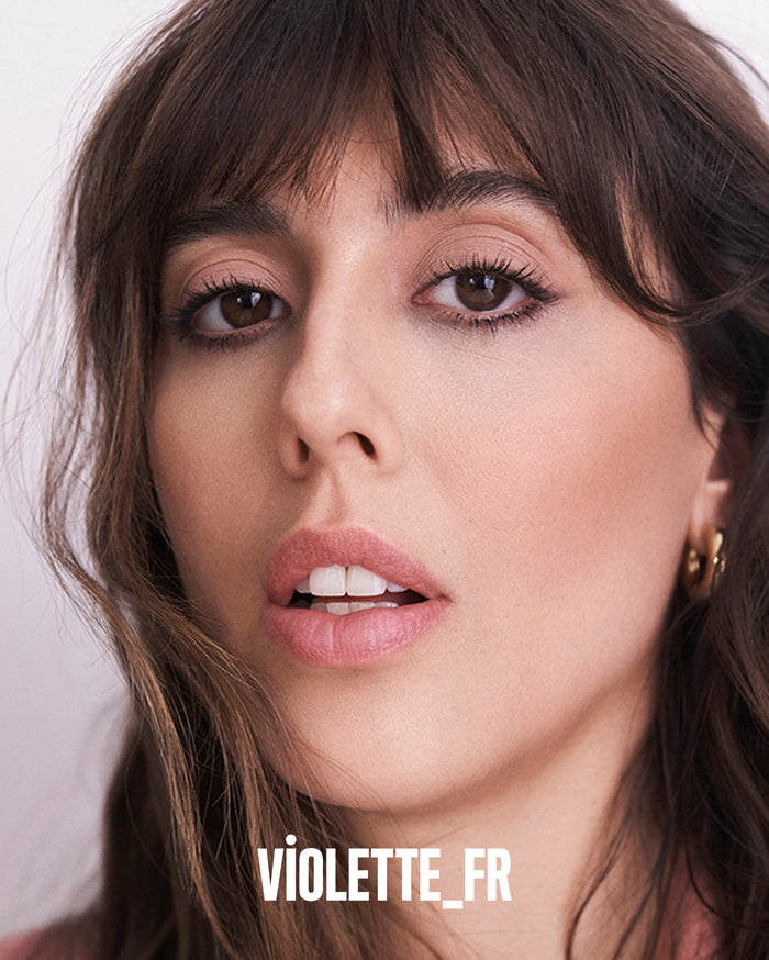 VIOLETTE_FR. Close-up of a woman with medium skin tone, dark brown hair and bangs, wearing natural eye makeup and pinkish-nude lipstick.