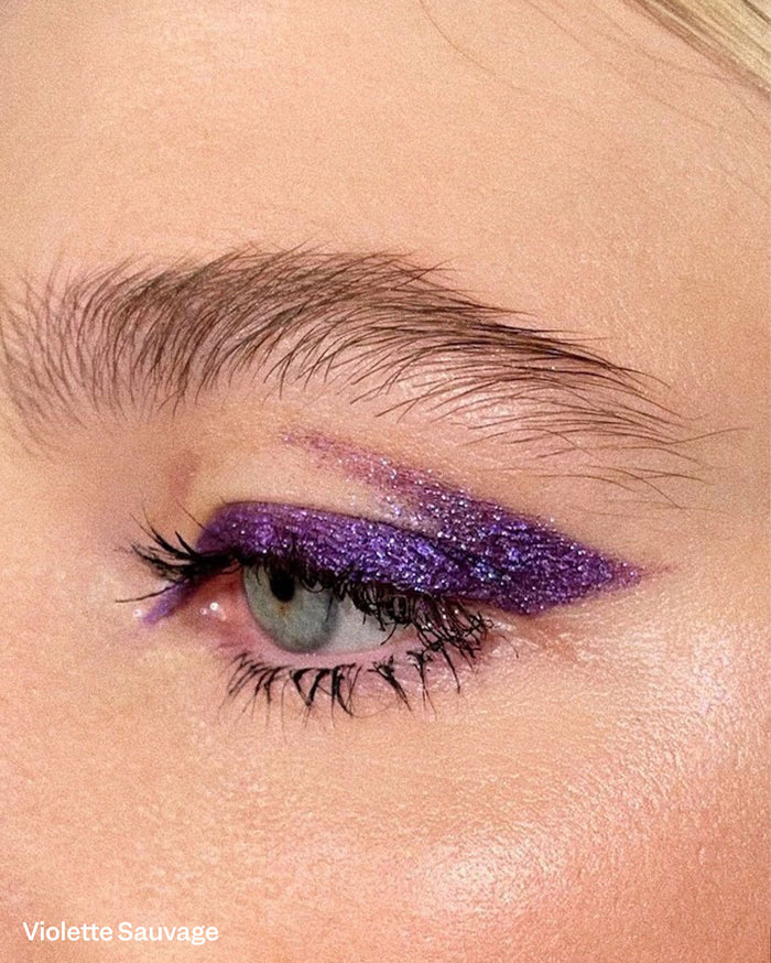 Violette Sauvage: Close-up of a light-skinned person's eye with a bold, glittery purple winged eyeliner and long dark lashes.