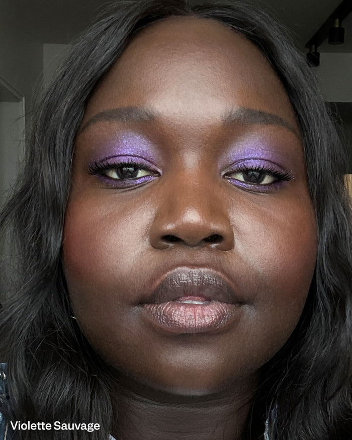 Violette Sauvage. Close-up of a model with deep skin tone wearing vibrant shimmery purple eyeshadow and a metallic plum lipstick.