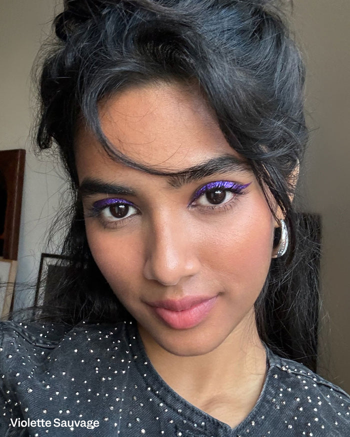 Violette Sauvage. A woman with deep skin tone models vibrant purple winged eyeliner and a natural pink lip.