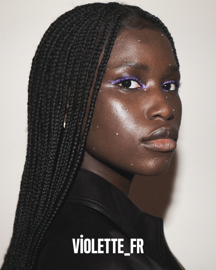 VIOLETTE_FR. Close-up of a model with deep skin tone, wearing vibrant purple glitter eyeliner and scattered rhinestones on her face, with long black box braids.